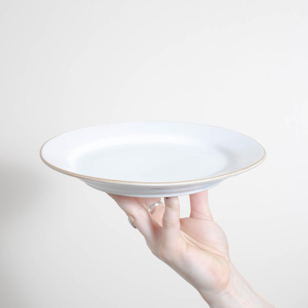 A hand holding a Bespoke 77 Matte White With Rye Edge 22.5cm High Rim Plate on a white background for food photography.