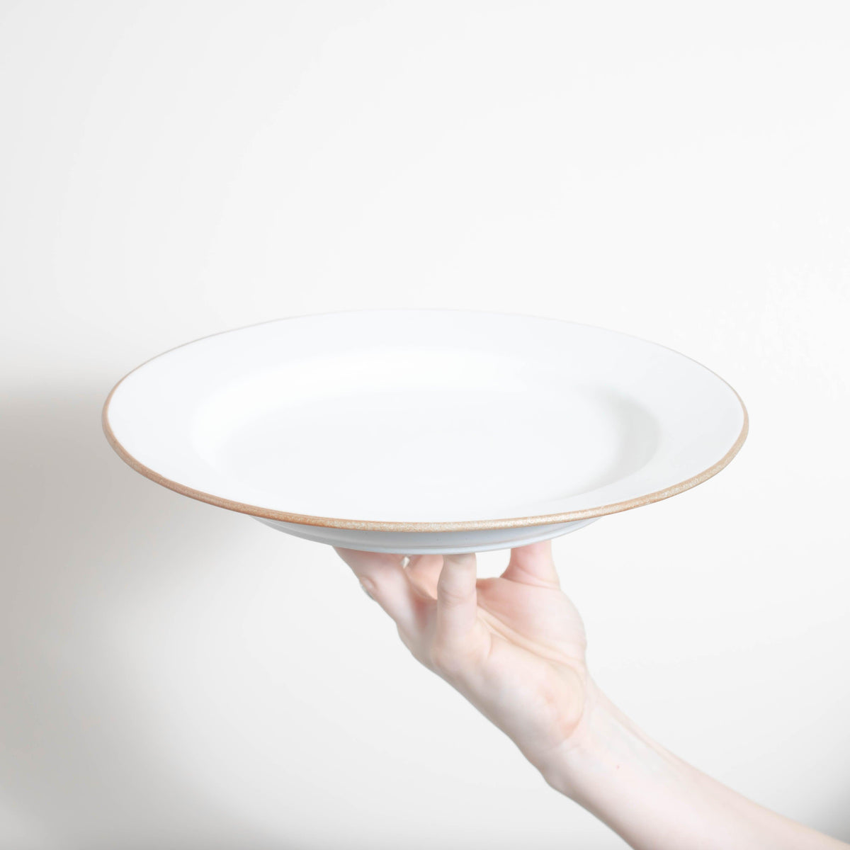 A person holding up a Matte White With Rye Edge 29cm Round Mains Plate by Bespoke 77, with a gold rim.